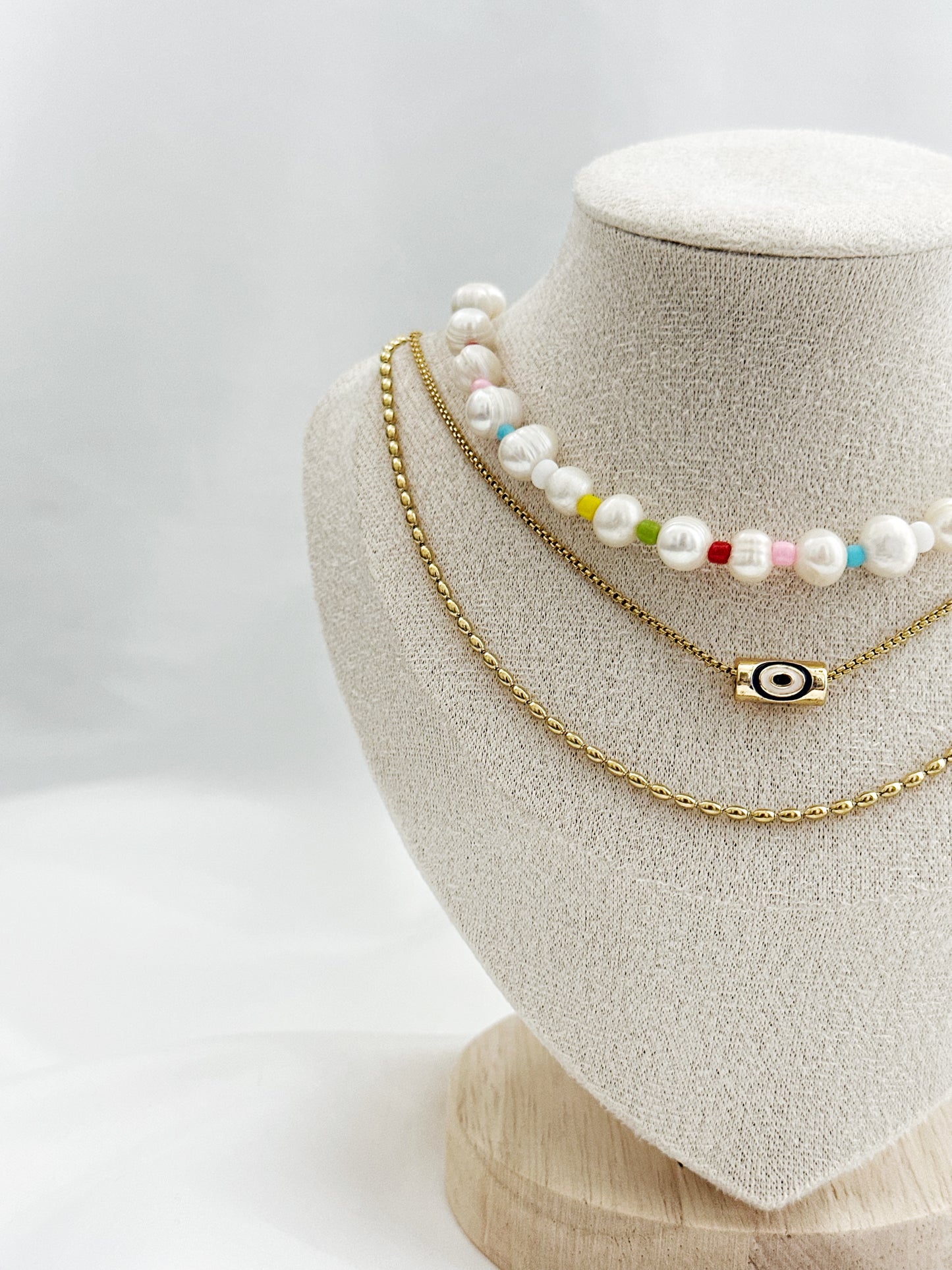 3-lagiges Kettenset mit unregelmäßigen Süßwasserperlen, bunter Perlenkette, Nazar-Anhänger und goldener Gliederkette – flach auf hellem Hintergrund arrangiert