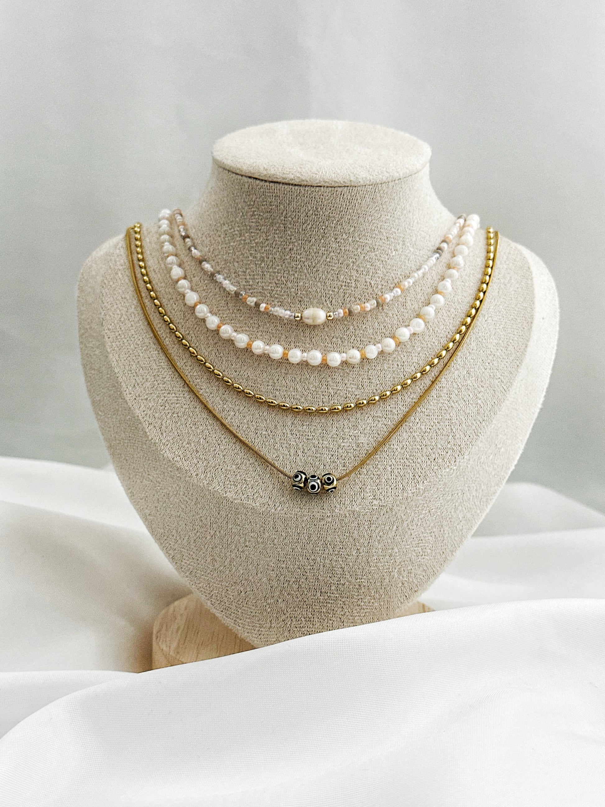 Layering-Ketten-Set bestehend aus Süßwasserperlen, pfirsichfarbenen Perlen, goldener Edelstahlkette und Nazar-Augen-Kette auf Samt-Büste fotografiert.