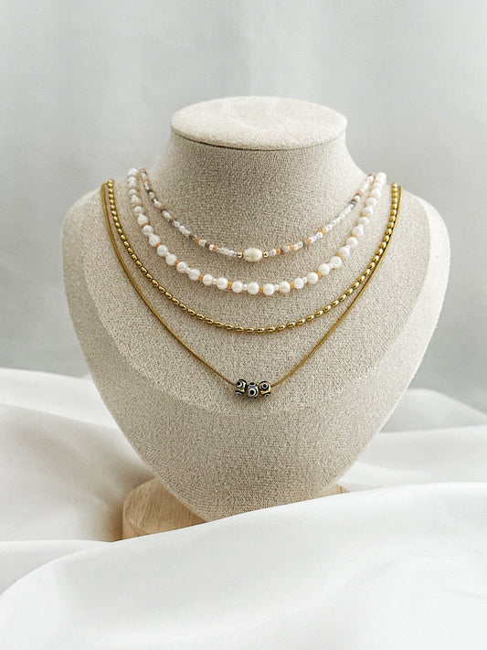 Layering-Ketten-Set bestehend aus Süßwasserperlen, pfirsichfarbenen Perlen, goldener Edelstahlkette und Nazar-Augen-Kette auf Samt-Büste fotografiert.