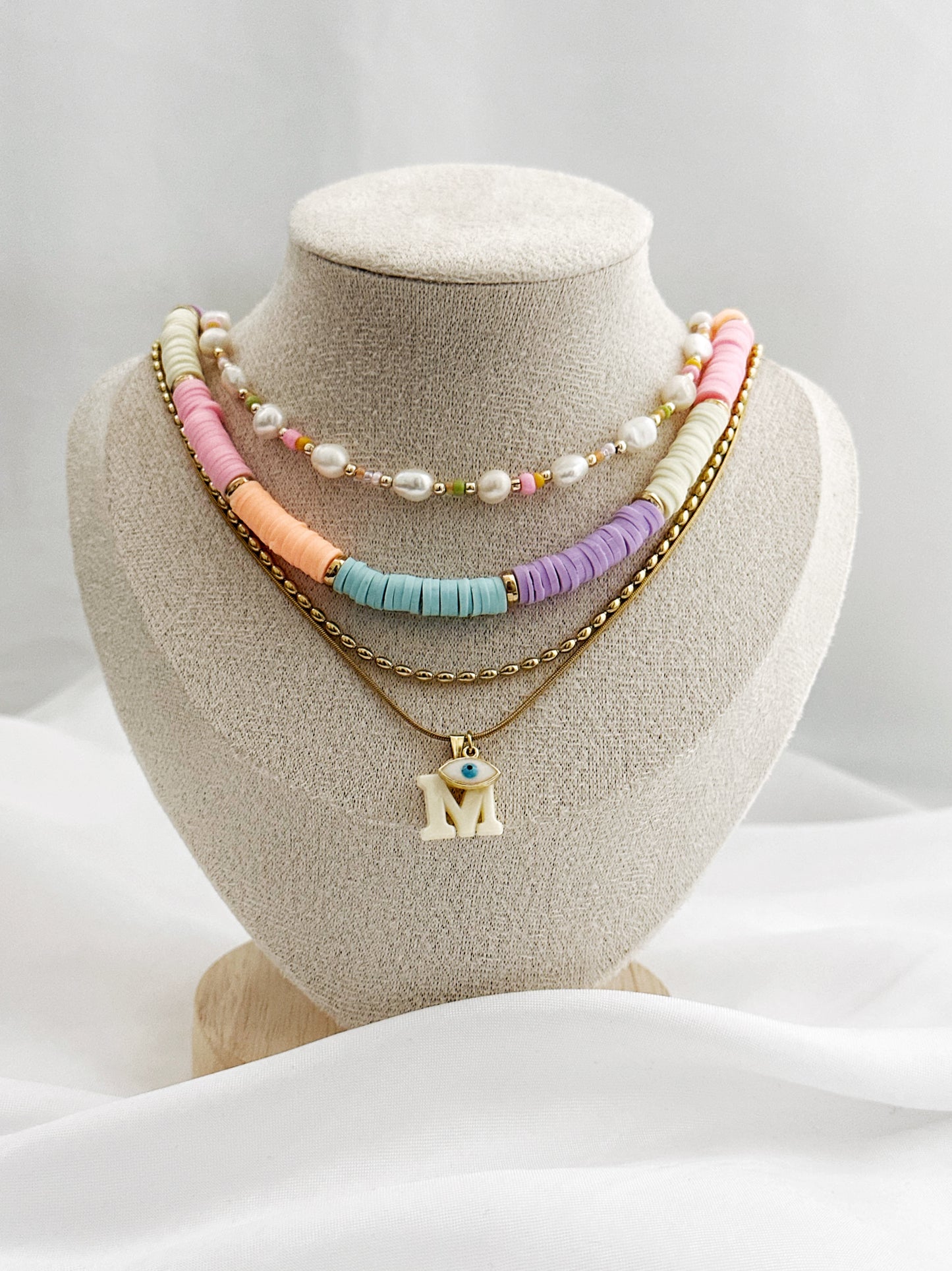 Layering-Set mit vier Halsketten auf Schmuckständer: Süßwasserperlenkette, bunte Polymerperlenkette, goldene Edelstahlkette und Initial-Anhänger mit Nazar-Auge