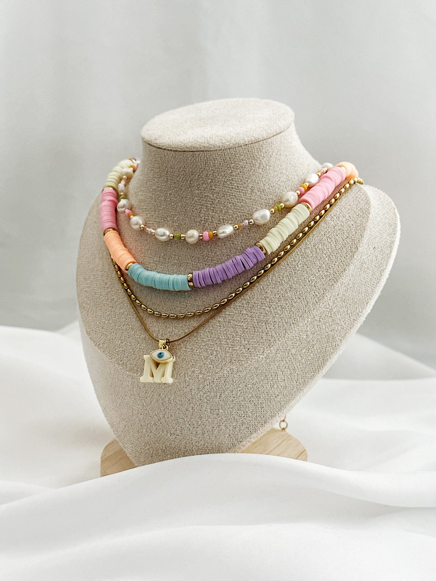 Layering-Set mit vier Halsketten auf Schmuckständer: Süßwasserperlenkette, bunte Polymerperlenkette, goldene Edelstahlkette und Initial-Anhänger mit Nazar-Auge