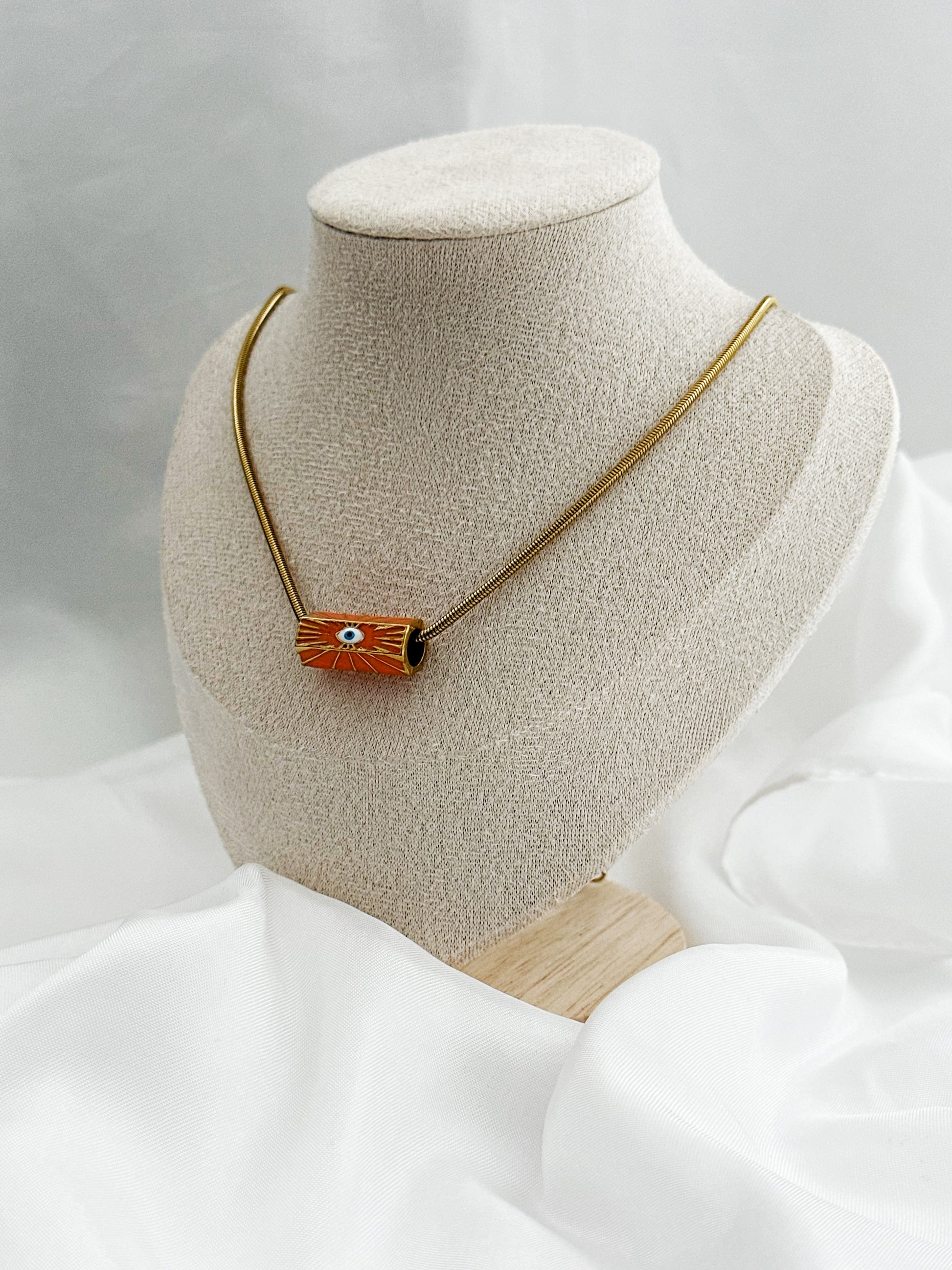 Goldene Edelstahl-Schlangenkette mit rundem orangefarbenem Anhänger – in der Mitte ein stilisiertes weißes Auge als Schutzsymbol 