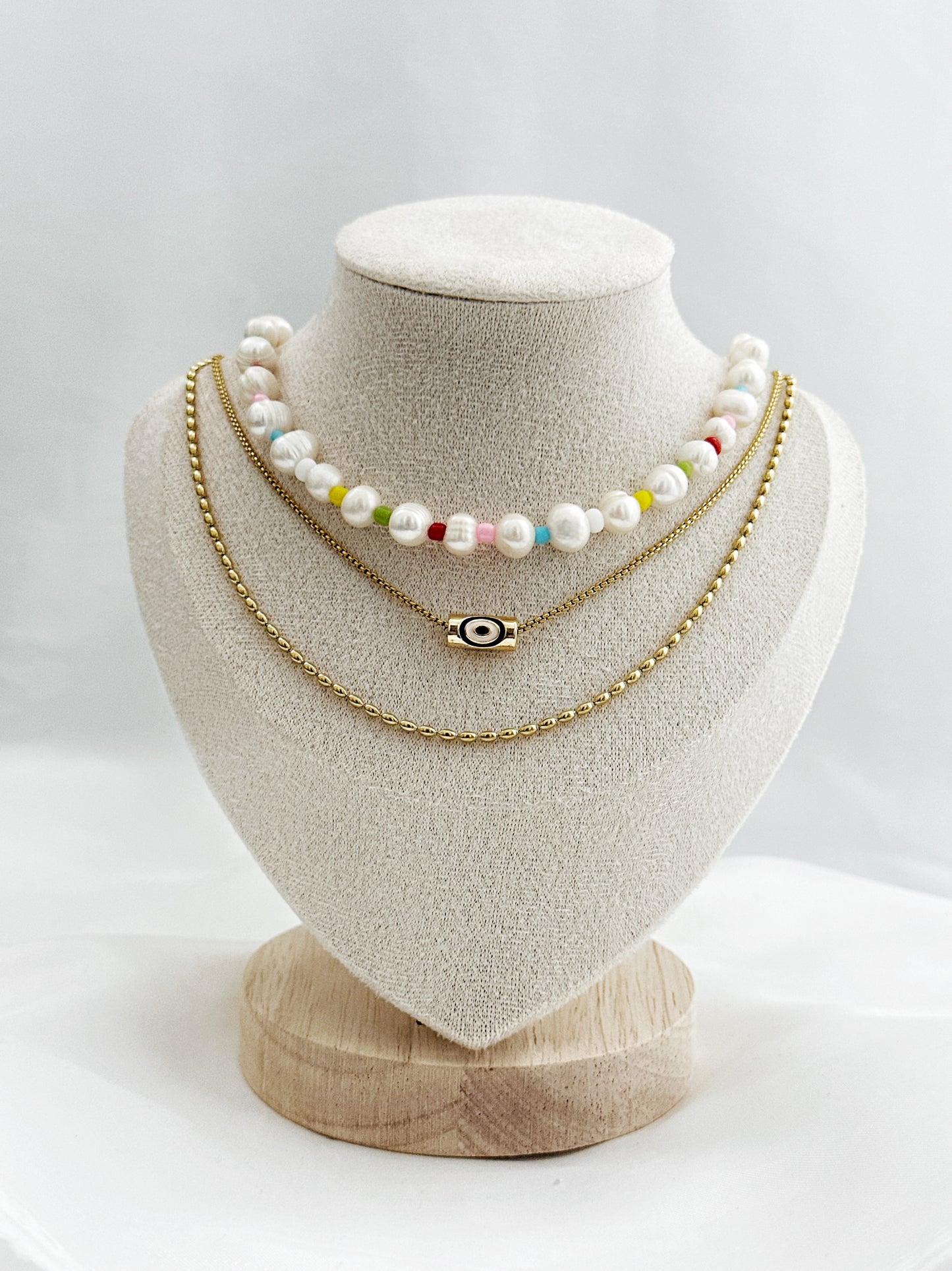 3-lagiges Kettenset mit unregelmäßigen Süßwasserperlen, bunter Perlenkette, Nazar-Anhänger und goldener Gliederkette – flach auf hellem Hintergrund arrangiert