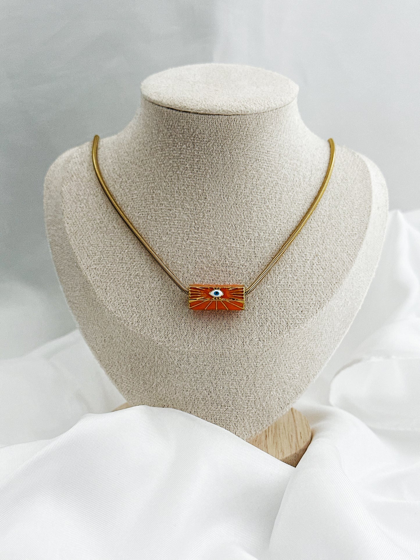 Goldene Edelstahl-Schlangenkette mit rundem orangefarbenem Anhänger – in der Mitte ein stilisiertes weißes Auge als Schutzsymbol 