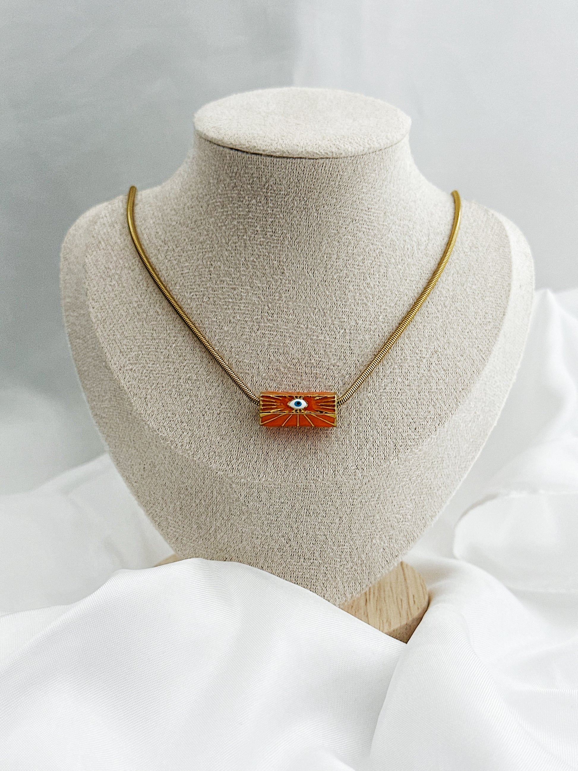 Goldene Edelstahl-Schlangenkette mit rundem orangefarbenem Anhänger – in der Mitte ein stilisiertes weißes Auge als Schutzsymbol 