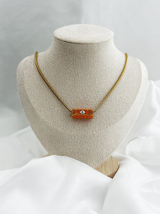 Goldene Edelstahl-Schlangenkette mit rundem orangefarbenem Anhänger – in der Mitte ein stilisiertes weißes Auge als Schutzsymbol 