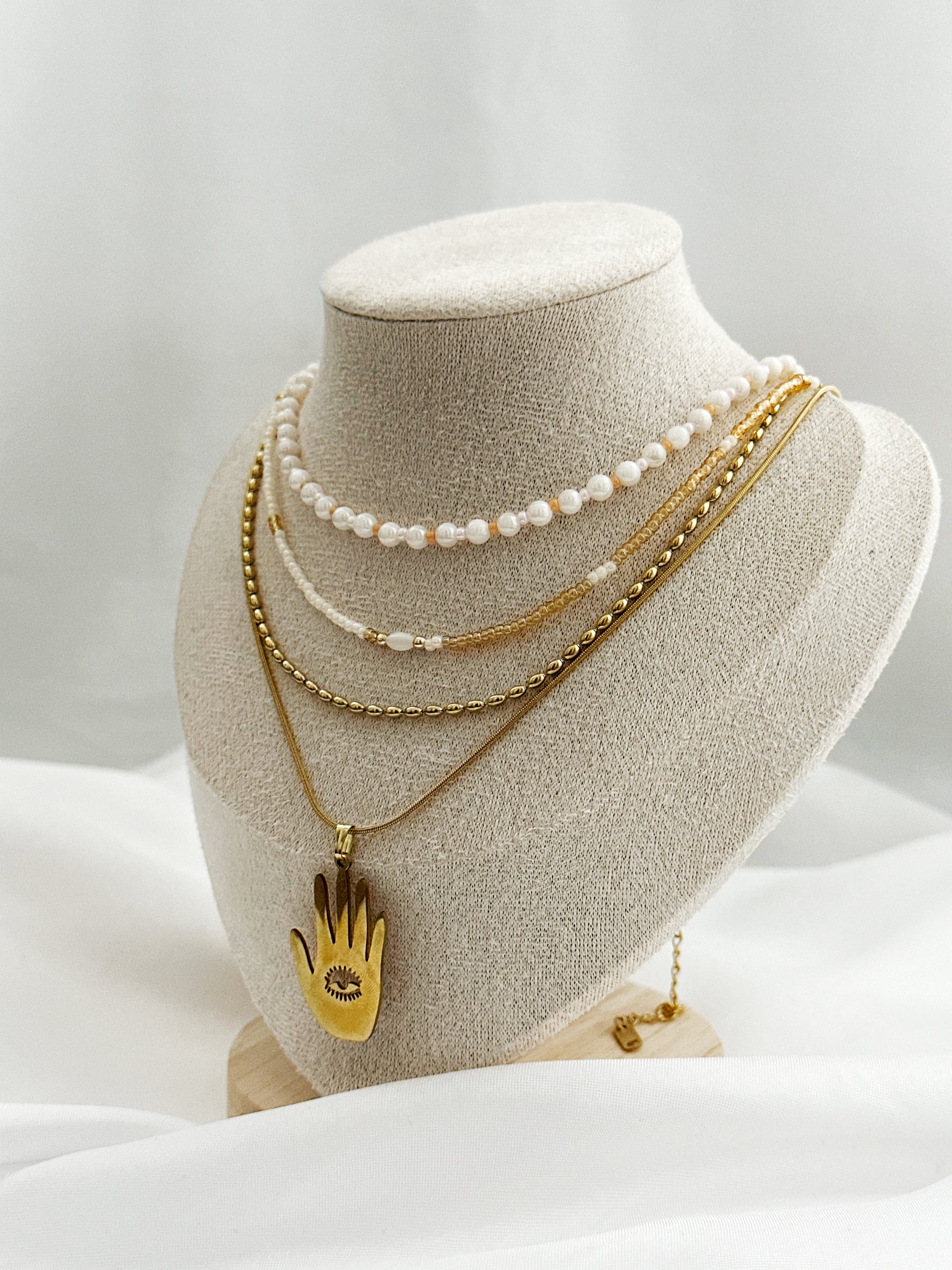 Elegantes Layering-Ketten-Set mit Hamsa-Anhänger, Süßwasserperlen und goldfarbenen Ketten auf Schmuckständer – 4-lagiger Schmucklook für Damen.