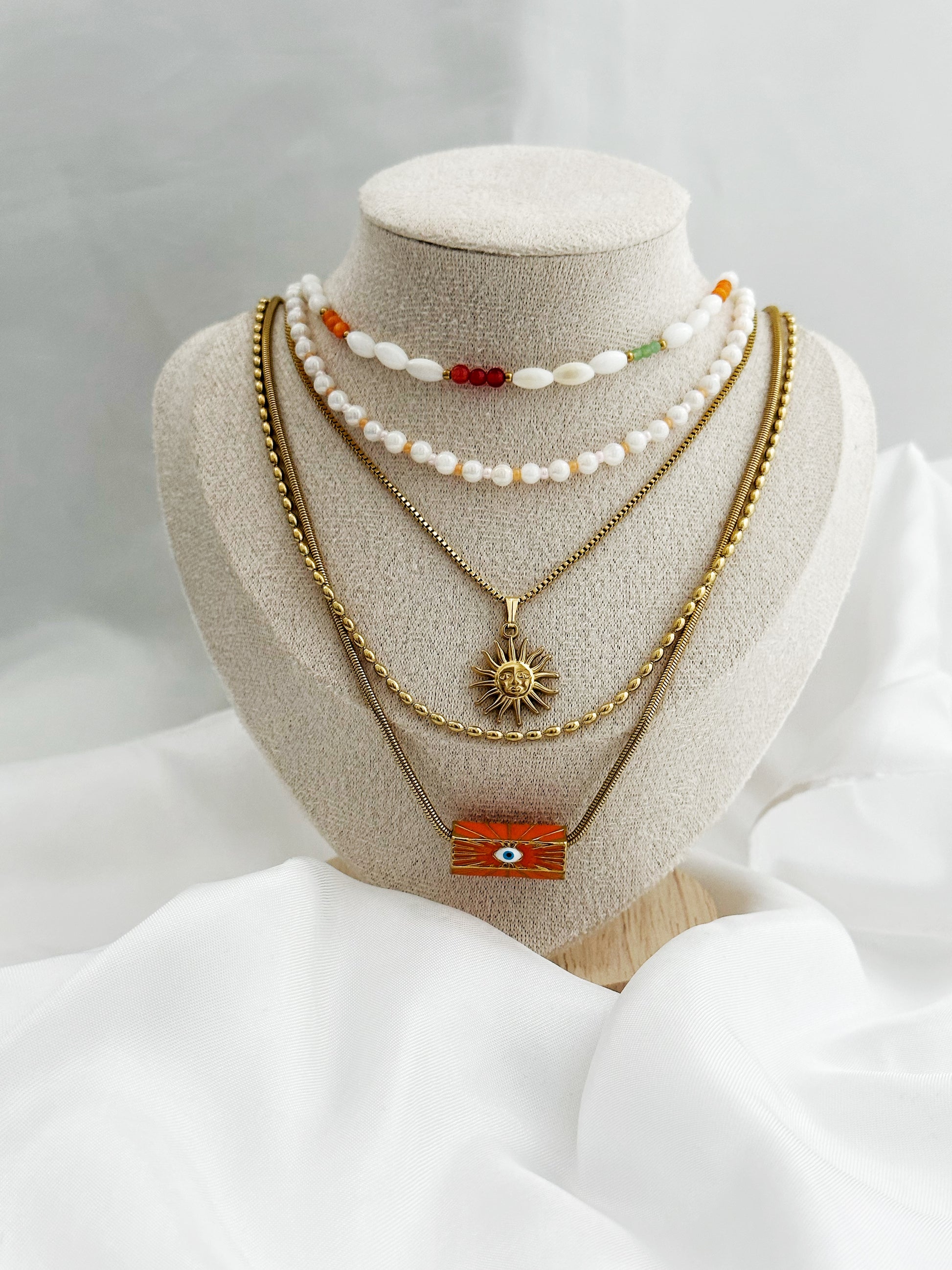 Layering-Ketten-Set mit fünf goldfarbenen und perlenbesetzten Halsketten auf Schmuckständer. Mit Sonnen-Anhänger, Nazar-Auge und natürlichen Perlen in Weiß, Orange und Gold
