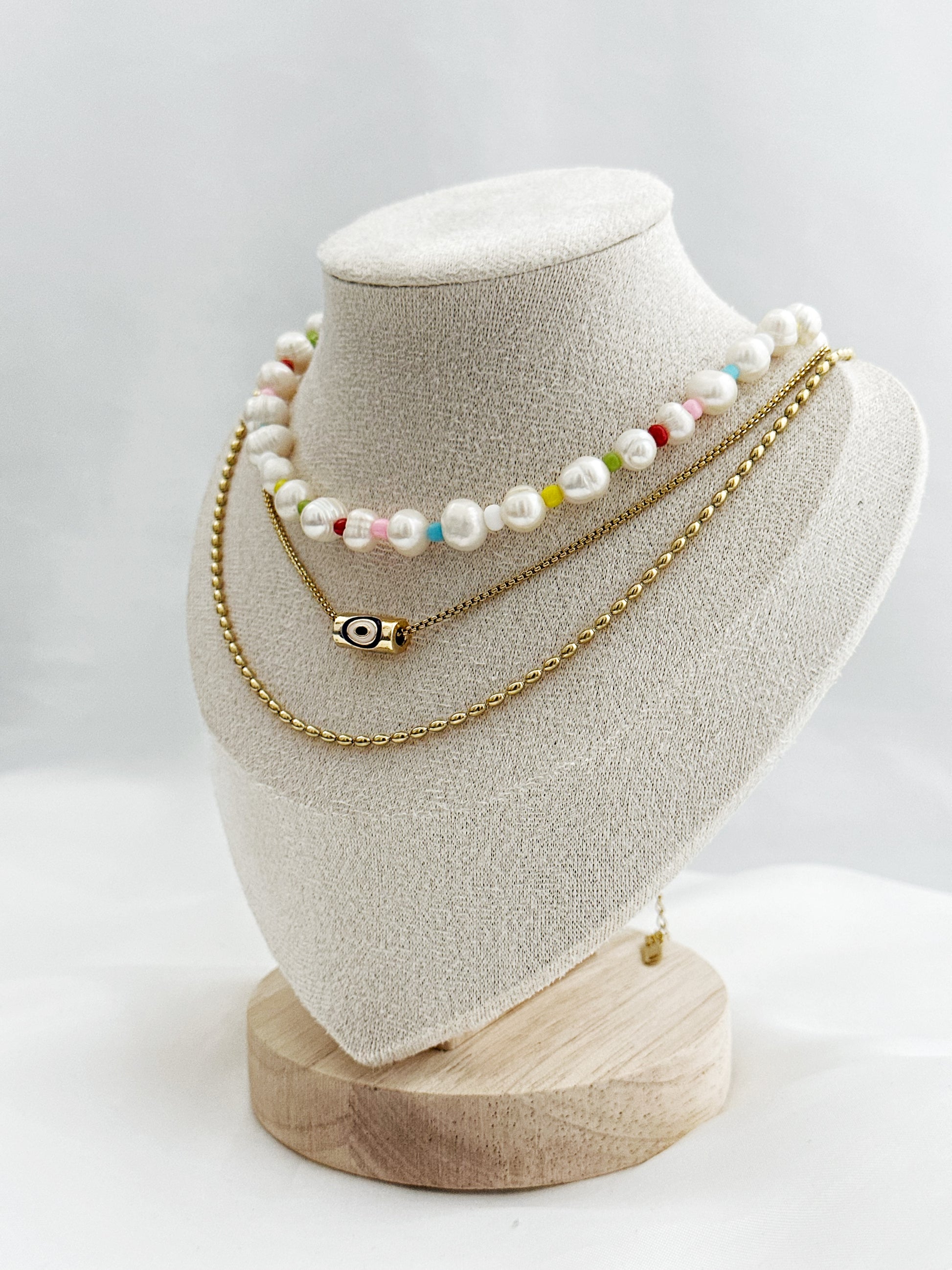 3-lagiges Kettenset mit unregelmäßigen Süßwasserperlen, bunter Perlenkette, Nazar-Anhänger und goldener Gliederkette – flach auf hellem Hintergrund arrangiert